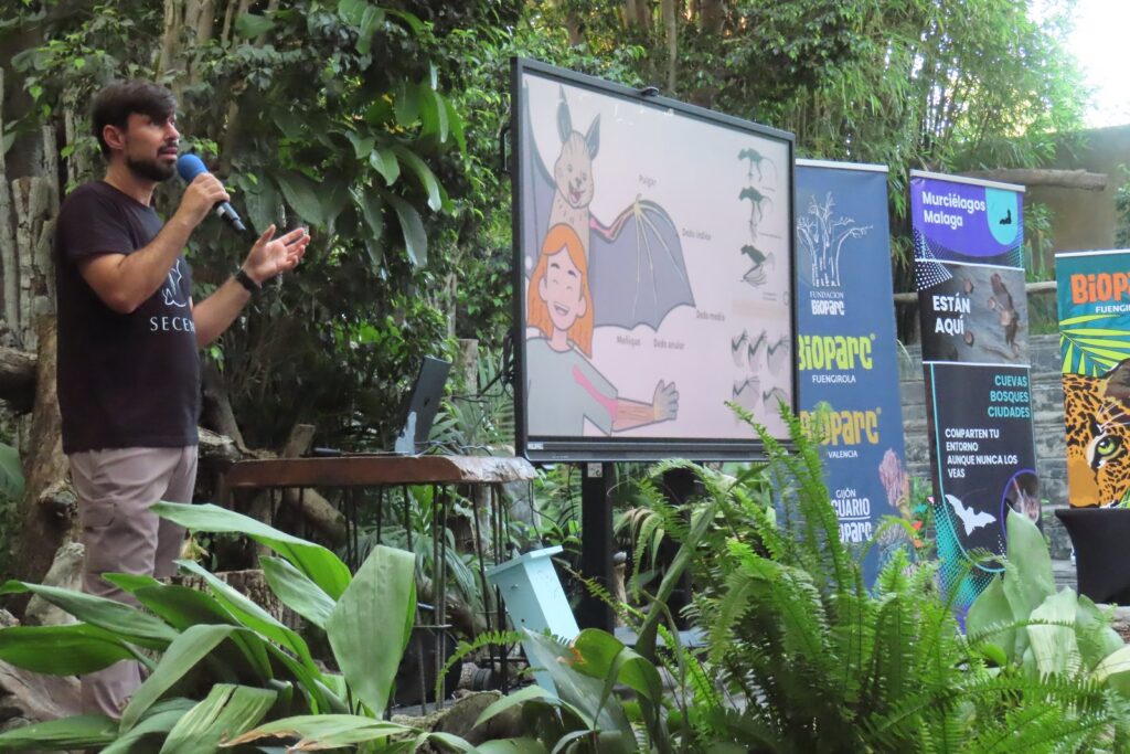 Charla de divulgación sobre murciélagos con micrófono, pantalla y paneles en un espacio ajardinado.